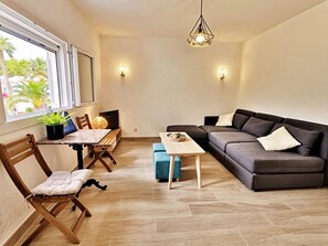 Ein Wohnzimmer mit Sofa, einem Esstisch mit Stühlen, einem Fernseher und einem Fenster mit Blick auf Palmen.