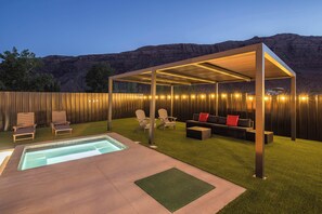 A covered outdoor seating area with a pool, chairs, and a pergola with string lights. - Luxury Moab Oasis Pool, Spa & Fire Pit (Moab)
