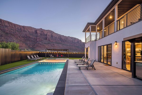 Un área de piscina moderna al aire libre con sillones de descanso, una zona de estar cubierta y vistas a una cordillera.