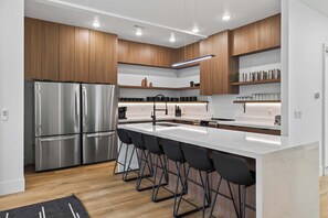 A modern kitchen with a stainless steel refrigerator, a central island with bar stools, wooden cabinets, and built-in shelves.