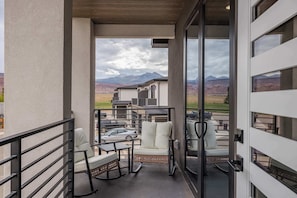 Un balcón con mobiliario exterior, vistas a montañas y un edificio moderno al fondo.
