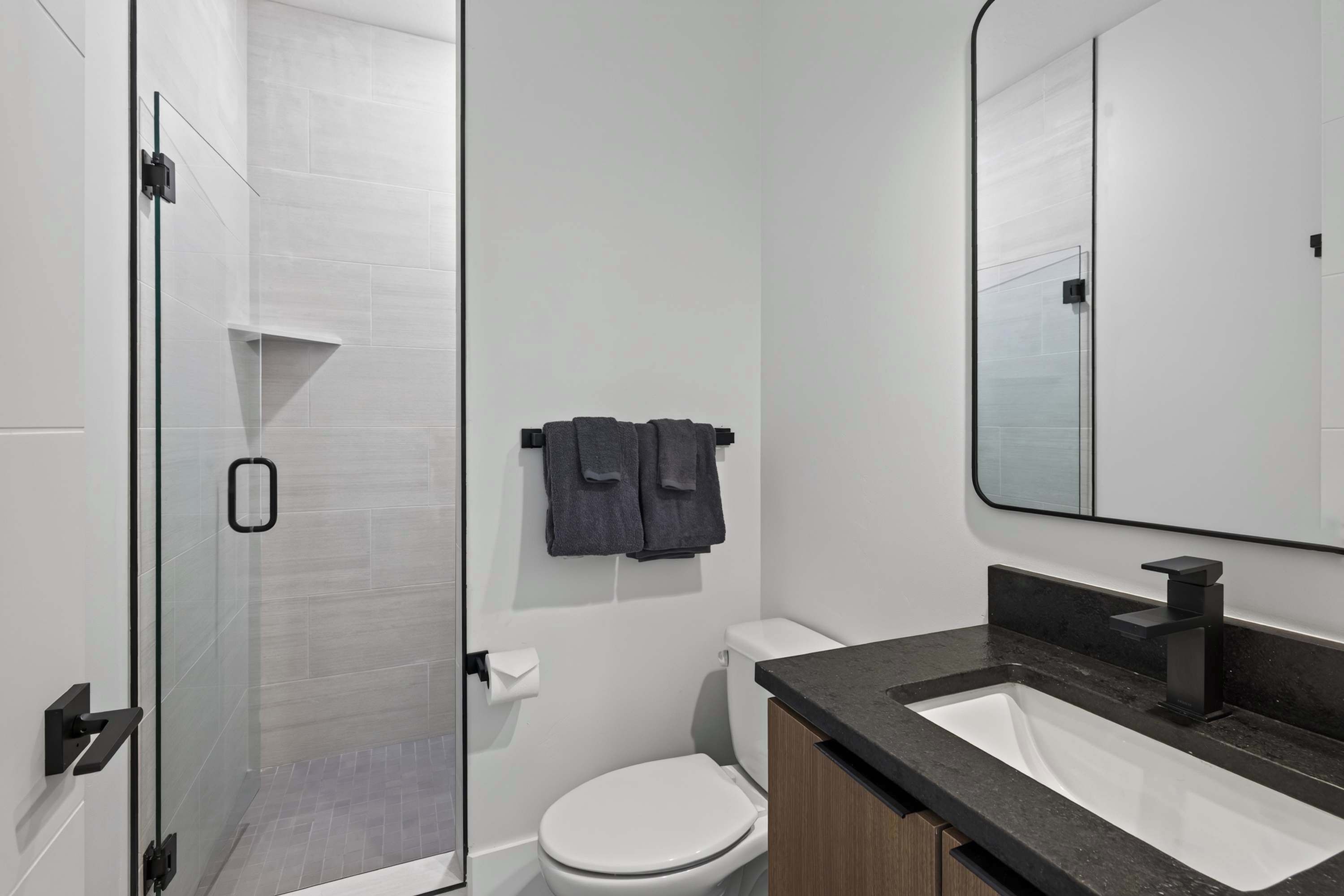 A modern bathroom with a glass shower enclosure, a white toilet, and a black sink with a wooden cabinet.