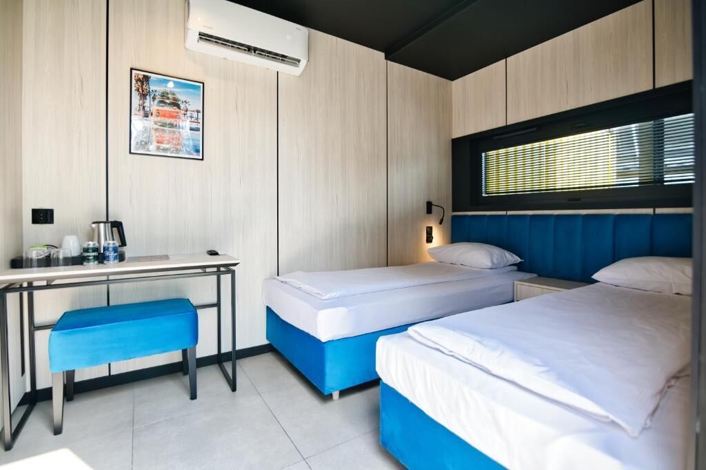 Habitación de hotel con escritorio, dos camas, una banca, aire acondicionado y ventana con persianas.