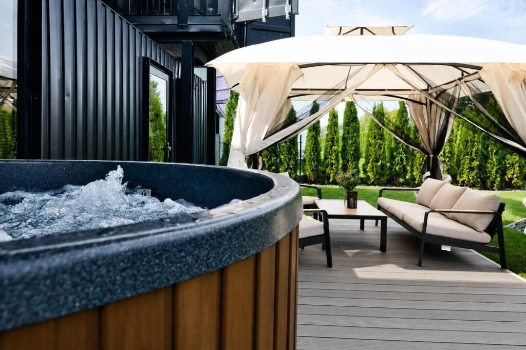 Ein Whirlpool auf einer Holzterrasse mit einer Couch und einem Tisch, umgeben von Grün.