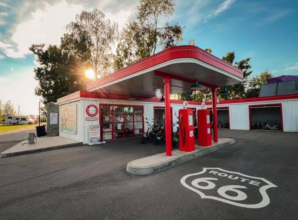 Um posto de gasolina com toldo vermelho, bombas de combustível e uma placa indicando a Rota 66.