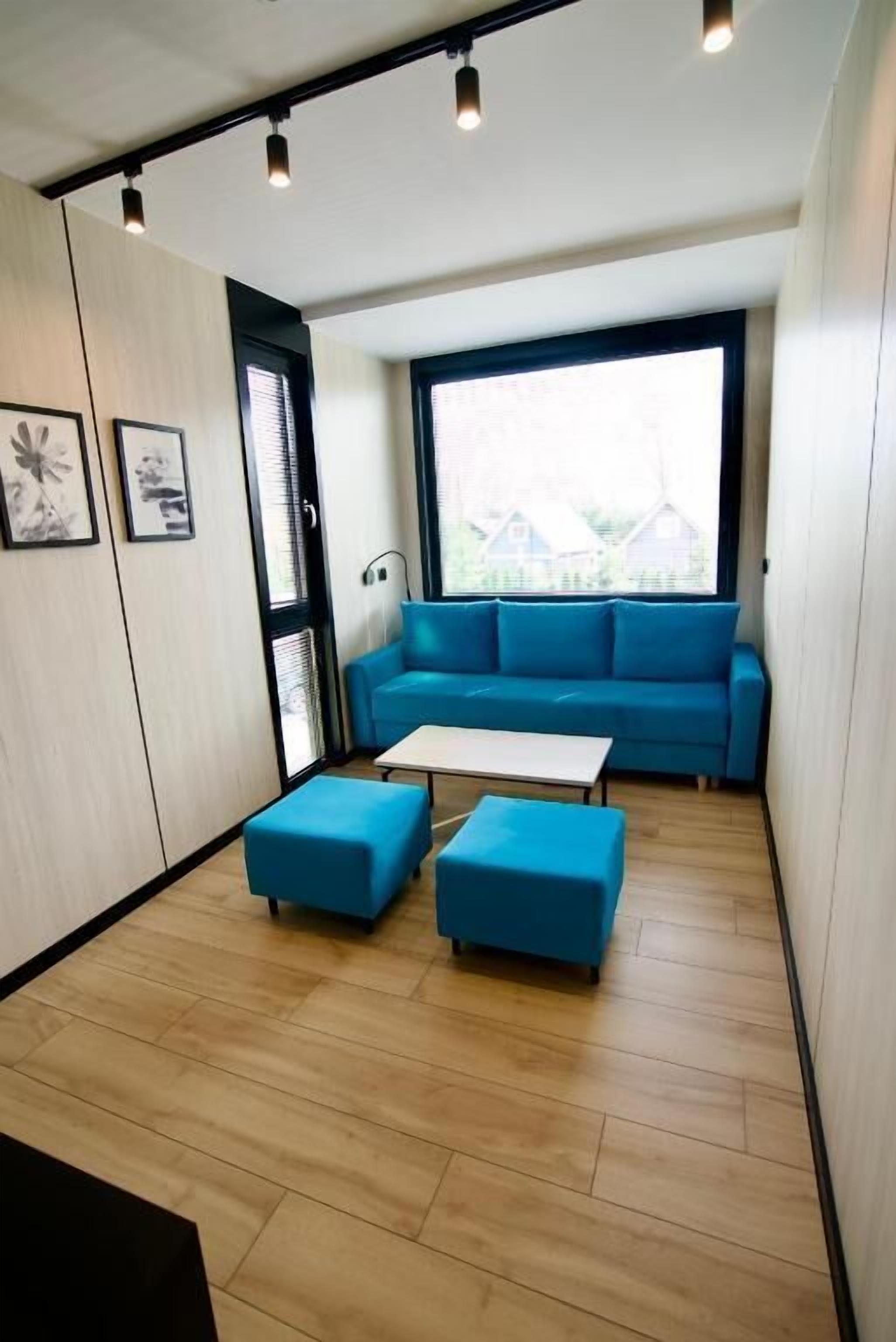 Una sala moderna con un sofá azul, dos otomanas a juego, una mesa de centro blanca y un ventanal con vistas al exterior.