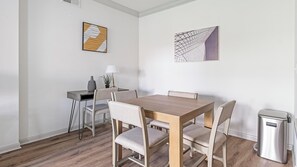 A dining area with a wooden table and chairs, a small desk with a lamp, and two wall art pieces. - Landing | Stunning 2BD, Gym, Clubhouse (Houston)