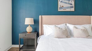 Une chambre à coucher avec un lit, des oreillers, une table de chevet, une lampe et une œuvre d’art murale.