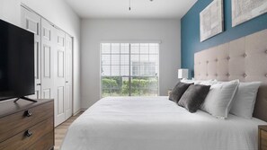 A bedroom with a bed, a television, a dresser, and a window with blinds. - Landing | Modern 2BD, Pool, Yoga Studio (Houston)