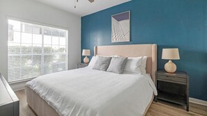 Ein Schlafzimmer mit einem Bett, zwei Nachttischen, einem Fenster mit Jalousien und einer Wand mit geometrischer Kunst.