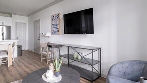 Uma sala de estar moderna com uma mesa de centro redonda, um sofá cinza, uma TV na parede e um espaço de jantar com mesa e cadeiras.