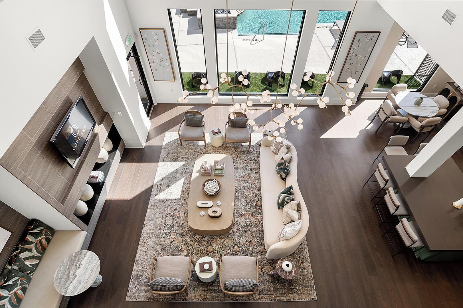 A modern living room with a large sofa, coffee table, and a TV. There is a pool visible through the windows.