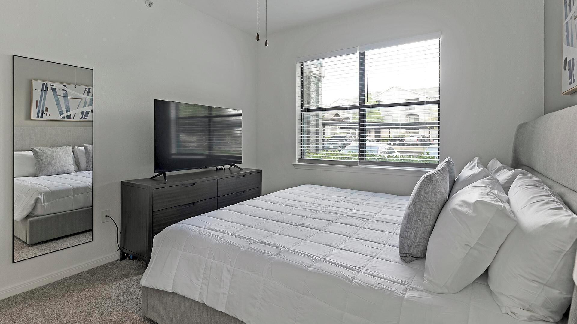 A bedroom with a bed, a dresser, a television, a window with blinds, and a wall-mounted mirror.