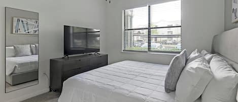 A bedroom with a bed, a dresser, a television, a window with blinds, and a wall-mounted mirror.