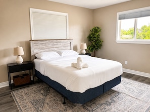 A neatly made bed with white linens, a dark blue woven bed skirt, and a matching blue headboard. There are two bedside tables with lamps, a potted plant, and a large window with blinds.