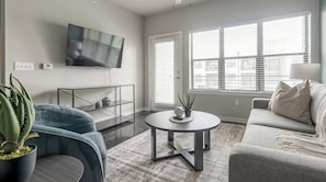 A modern living room with a flat-screen TV mounted on the wall, a round coffee table, a sofa, and a potted plant. - Landing | Stunning 1BD, Pool, Gym (Austin)