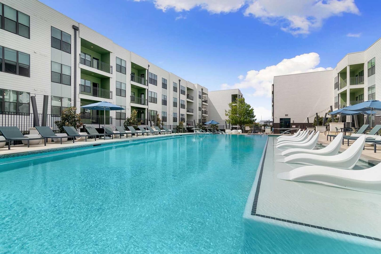 A swimming pool with lounge chairs and umbrellas, surrounded by multi-story buildings with balconies.