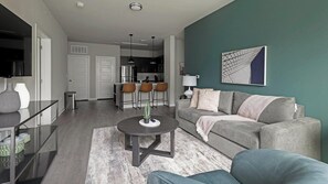 A modern living room with a grey sofa, a coffee table, and a TV. There is a kitchen in the background with a dining area. - Landing | Amazing 2BD, Pool, Gym (Austin)