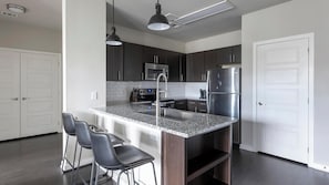A modern kitchen with dark cabinets, stainless steel appliances, a granite countertop, and a bar stool. - Landing | Spectacular 2BD, Pool, Gym (Austin)