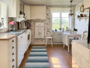 Une cuisine équipée d’un évier, d’une cuisinière et de placards, une fenêtre avec des rideaux et un petit tabouret.