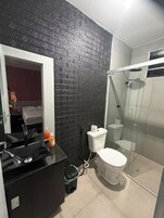 Casa de banho moderna com parede de tijolos pretos, cabine de chuveiro em vidro e bancada preta com lavatório e espelho.