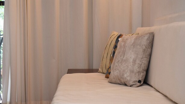 Una cama con colchón blanco y dos almohadas, una con estampado geométrico, en una habitación con cortinas traslúcidas.