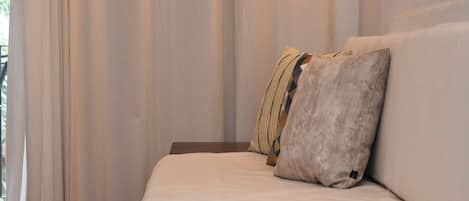 Una cama con colchón blanco y dos almohadas, una con estampado geométrico, en una habitación con cortinas traslúcidas.