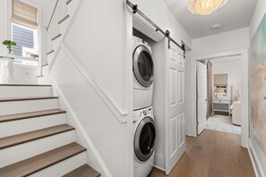 Una sala de lavandería con dos lavadoras y secadoras de carga frontal, una escalera de madera y un pasillo que conduce a un baño.