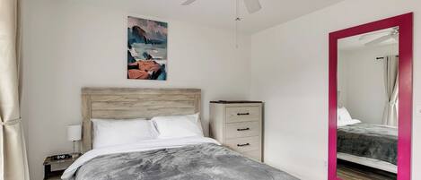 A bedroom with a bed, a nightstand, a dresser, a ceiling fan, a painting, and a mirror.