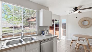 Una cocina con lavavajillas, un lavabo y una ventana con persianas. Hay un ventilador de techo y una zona de comedor con una mesa y sillas.