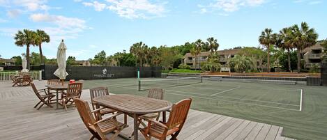 An outdoor tennis court with wooden deck seating and palm trees.