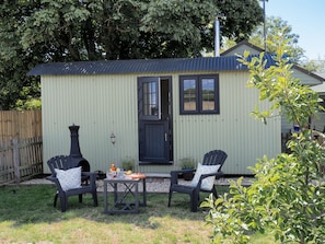 A small, single-story building with a corrugated metal roof, a door, and windows. There are two chairs and a small table with items on it.