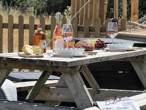 A wooden picnic table set outdoors with food, drinks, and a bottle.