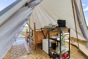 A tent interior with a bed, a table with a kettle, and a small refrigerator.