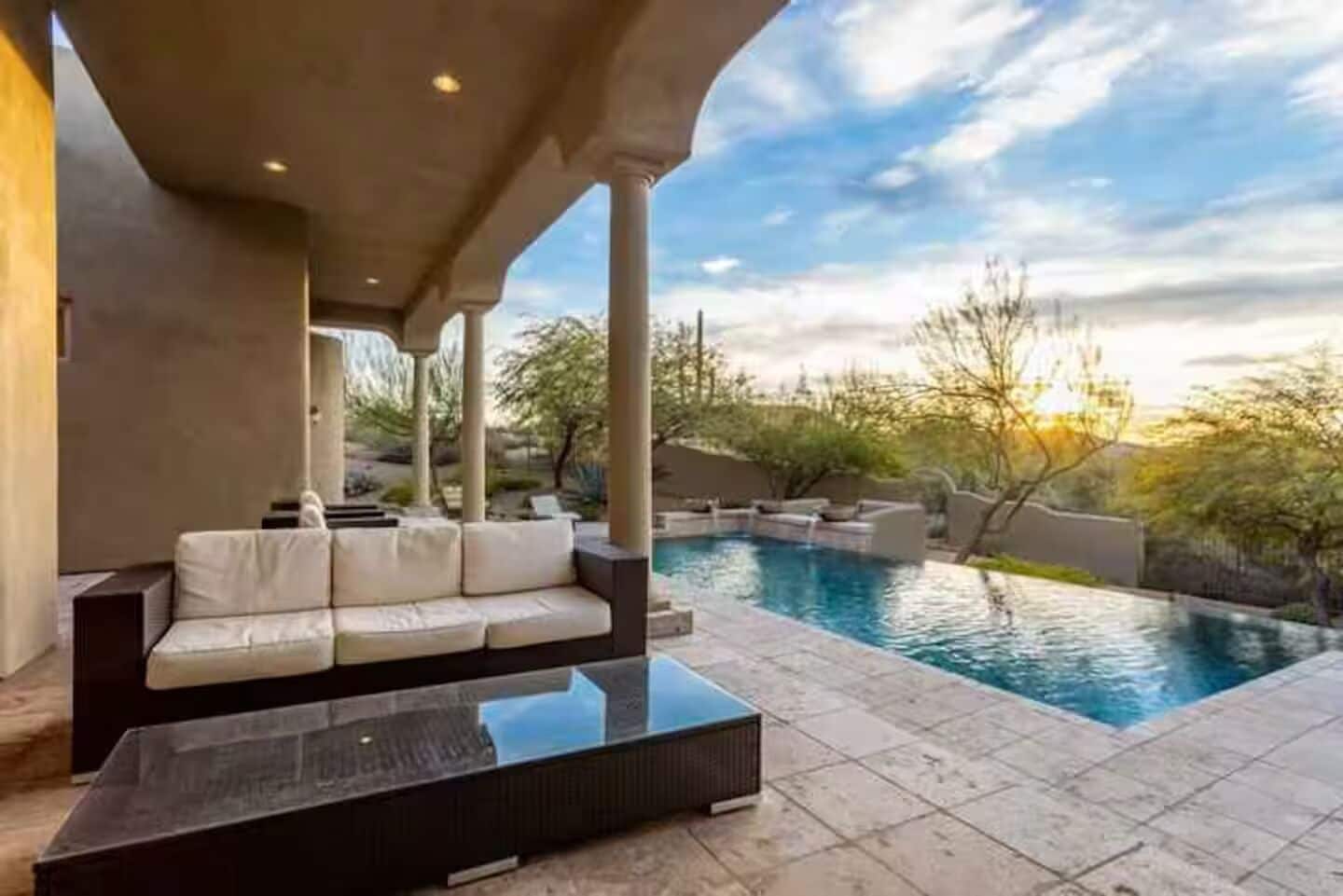 A pool area with a sofa and a coffee table, surrounded by a covered patio.