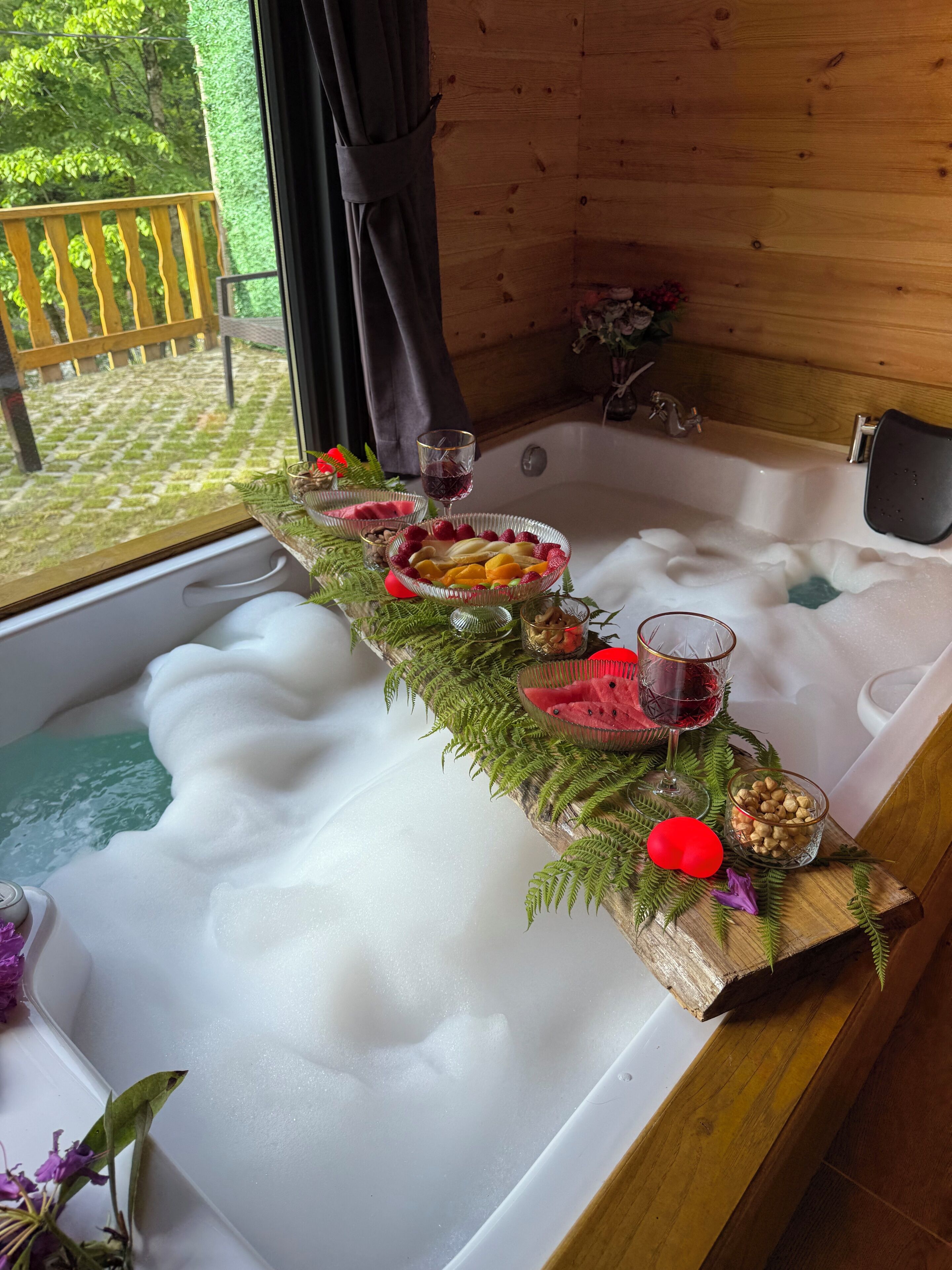 A wooden cabin with a bathtub filled with bubbles, surrounded by various fruits, nuts, and a glass of red wine.