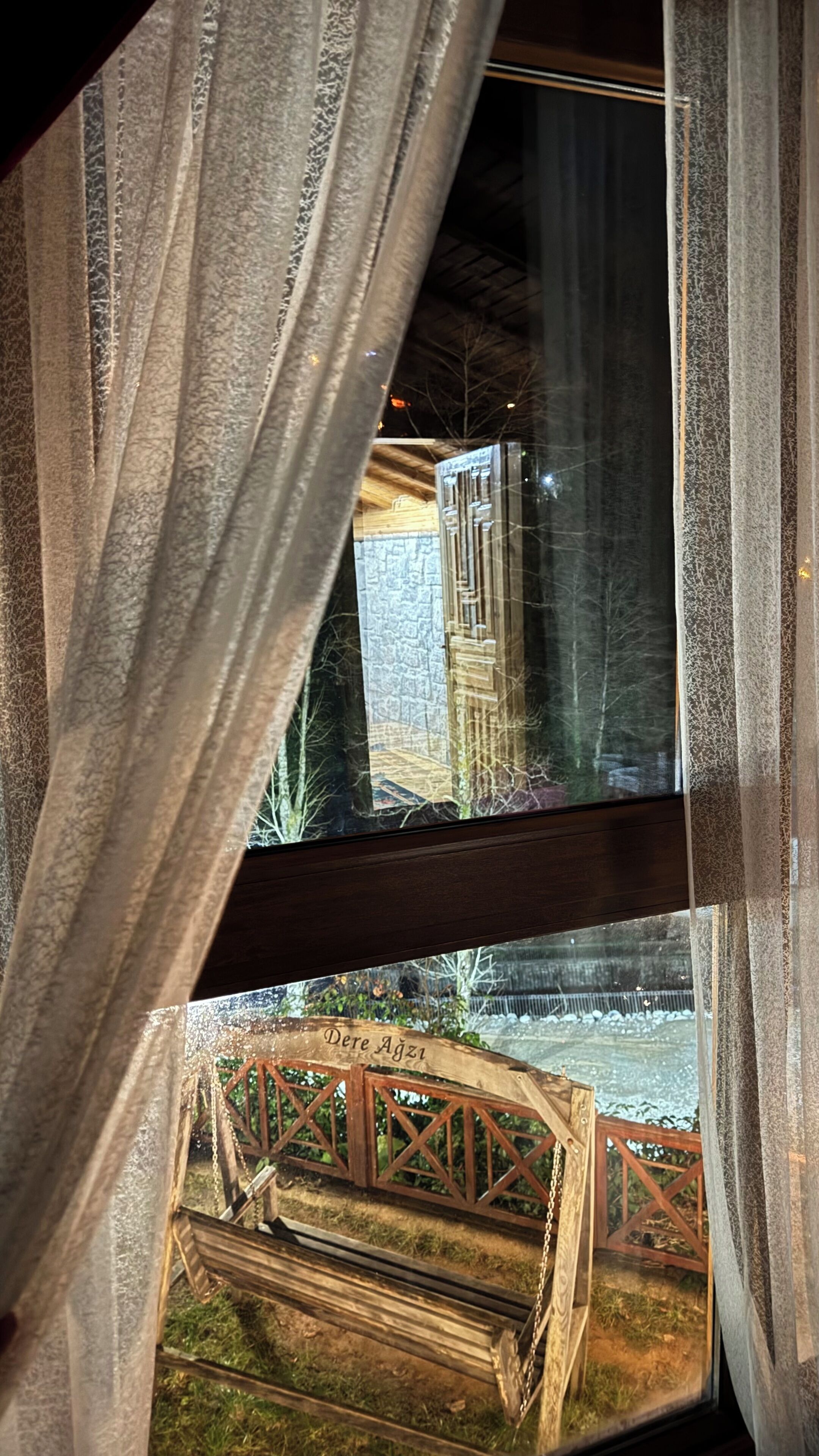 Vista a través de una ventana que muestra una estructura de madera en el exterior, posiblemente un puente o una puerta, y un muro de piedra al fondo.