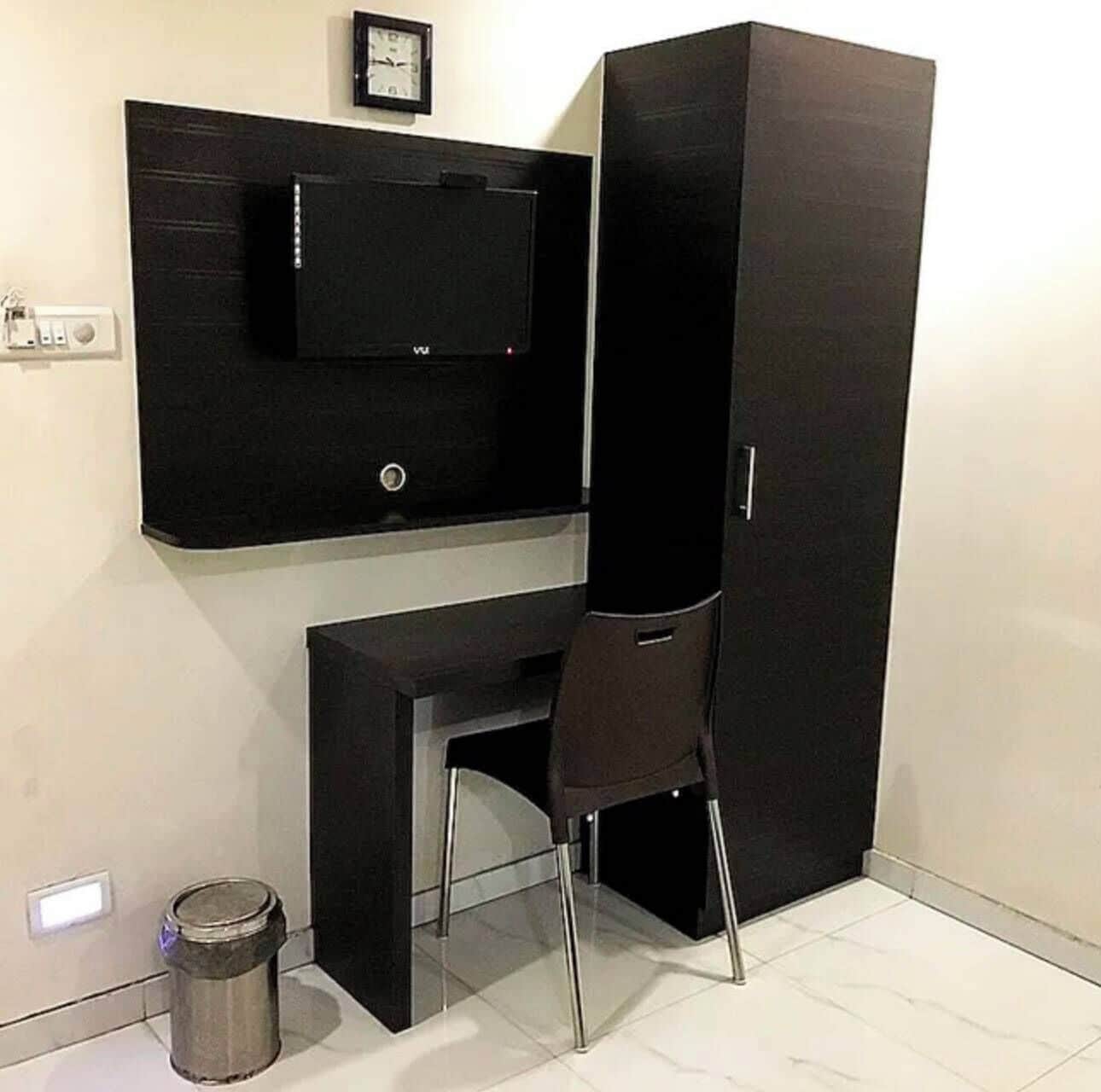 A compact room with a black desk, a chair, a television mounted on the wall, and a trash bin.