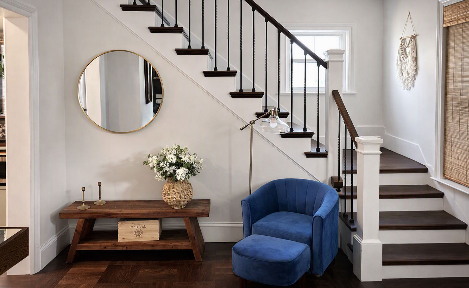 A staircase with dark wooden steps and black balusters, a round mirror on the wall, a blue armchair, a wicker basket with flowers, and a floor lamp.