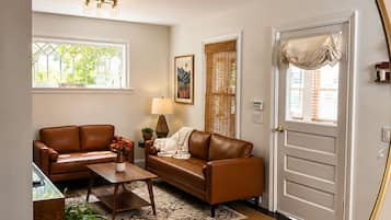 A living room with a brown leather sofa, a coffee table, and a lamp. There is a window with a view of greenery outside.