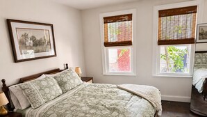 Un dormitorio con cama, mesitas de noche, un cuadro enmarco y ventanas con persianas.