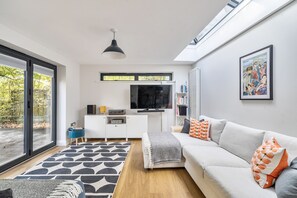 A modern living room with a sofa, a TV, a bookshelf, and a large window. - Coach House Retreat - Beaminster, Dorset (Beaminster)