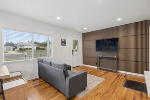 A modern living room with a grey sofa, wooden flooring, a TV on a wall, and a window with blinds. - Downtown 2 bedroom ranch home with hot tub, AC and all the amenities! (Colorado Springs)