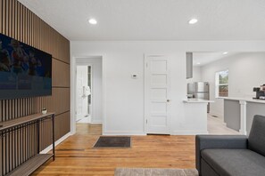 A modern living room with a grey sofa, wooden flooring, a white door, and a kitchen area with a fridge and cabinets. - Downtown 2 bedroom ranch home with hot tub, AC and all the amenities! (Colorado Springs)