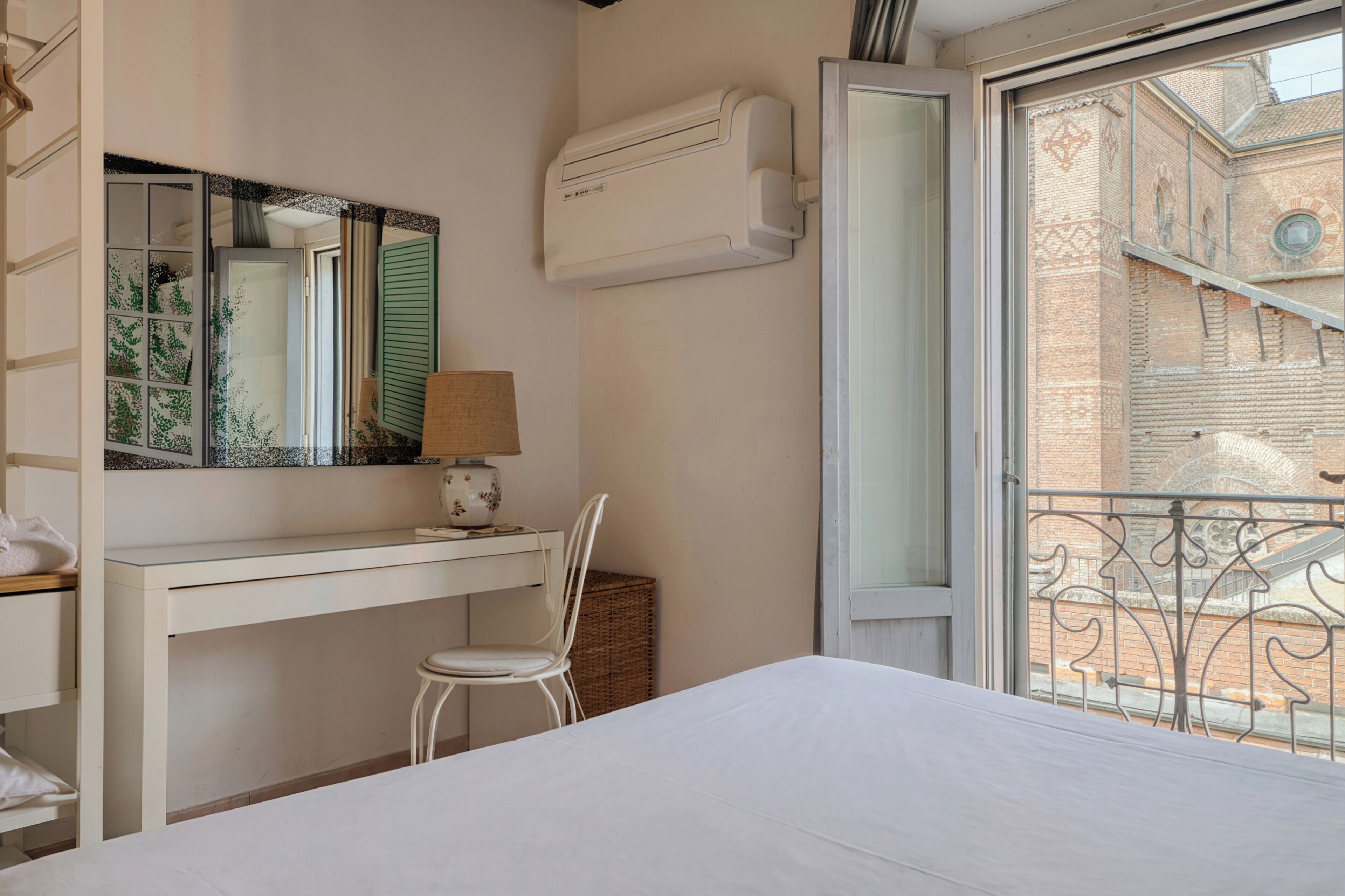 Un dormitorio con cama, escritorio, silla, lámpara y ventana con vista a un edificio.