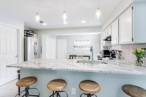 A modern kitchen with a marble countertop, bar stools, hanging pendant lights, and stainless steel appliances.
