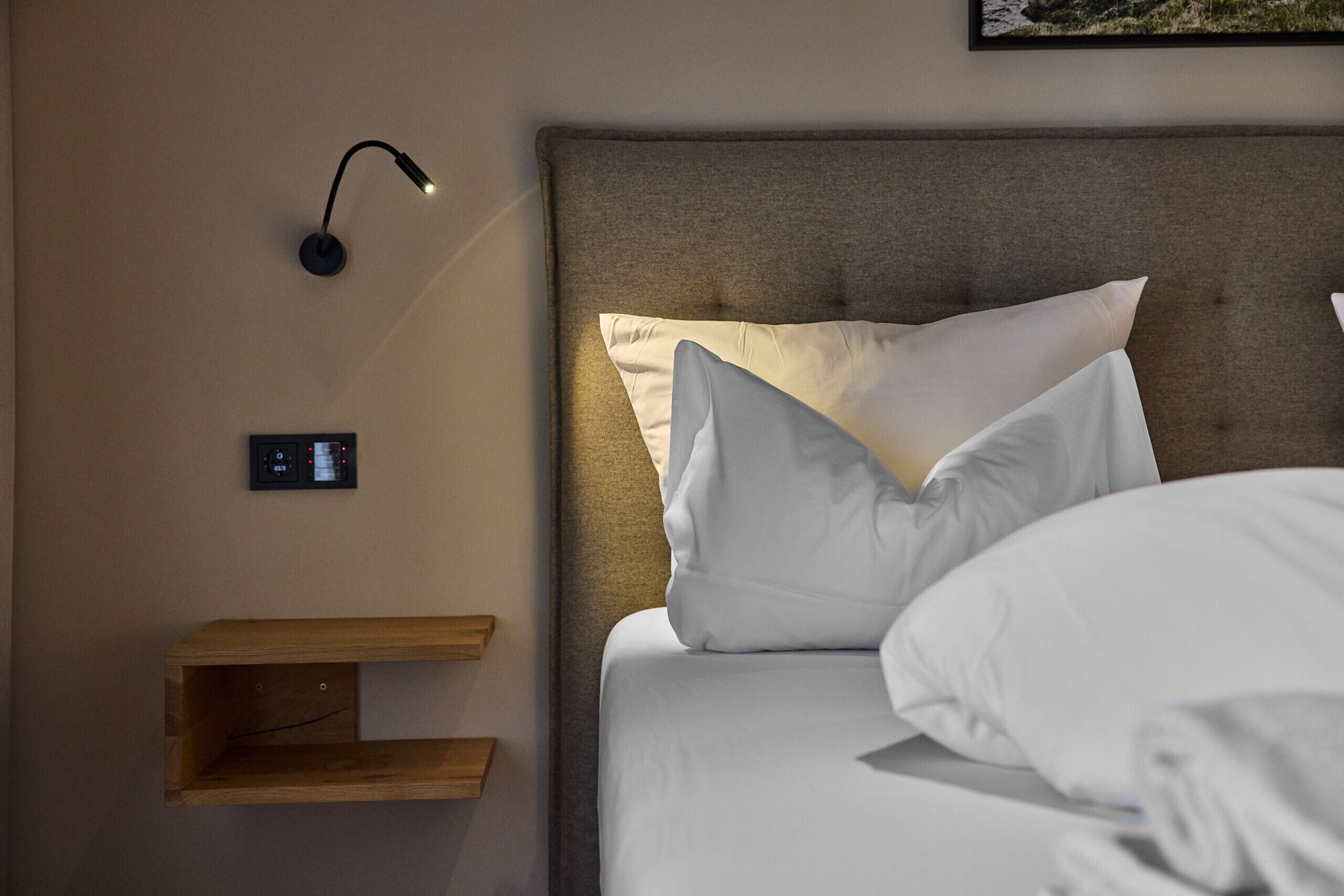 A neatly made bed with white linens and pillows, a bedside table with a lamp, and a wall-mounted control panel.