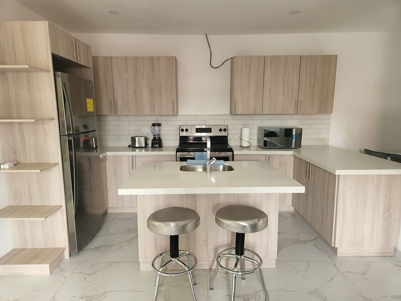 A modern kitchen with light wood cabinets, a stainless steel refrigerator, a double sink, and two bar stools.