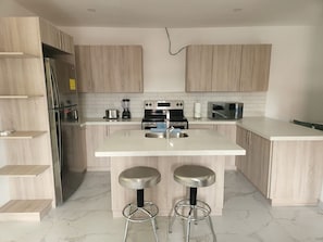 A modern kitchen with light wood cabinets, a stainless steel refrigerator, a double sink, and two bar stools. - Two bedroom  in Honduras, San Pedro Sula (San Pedro Sula)