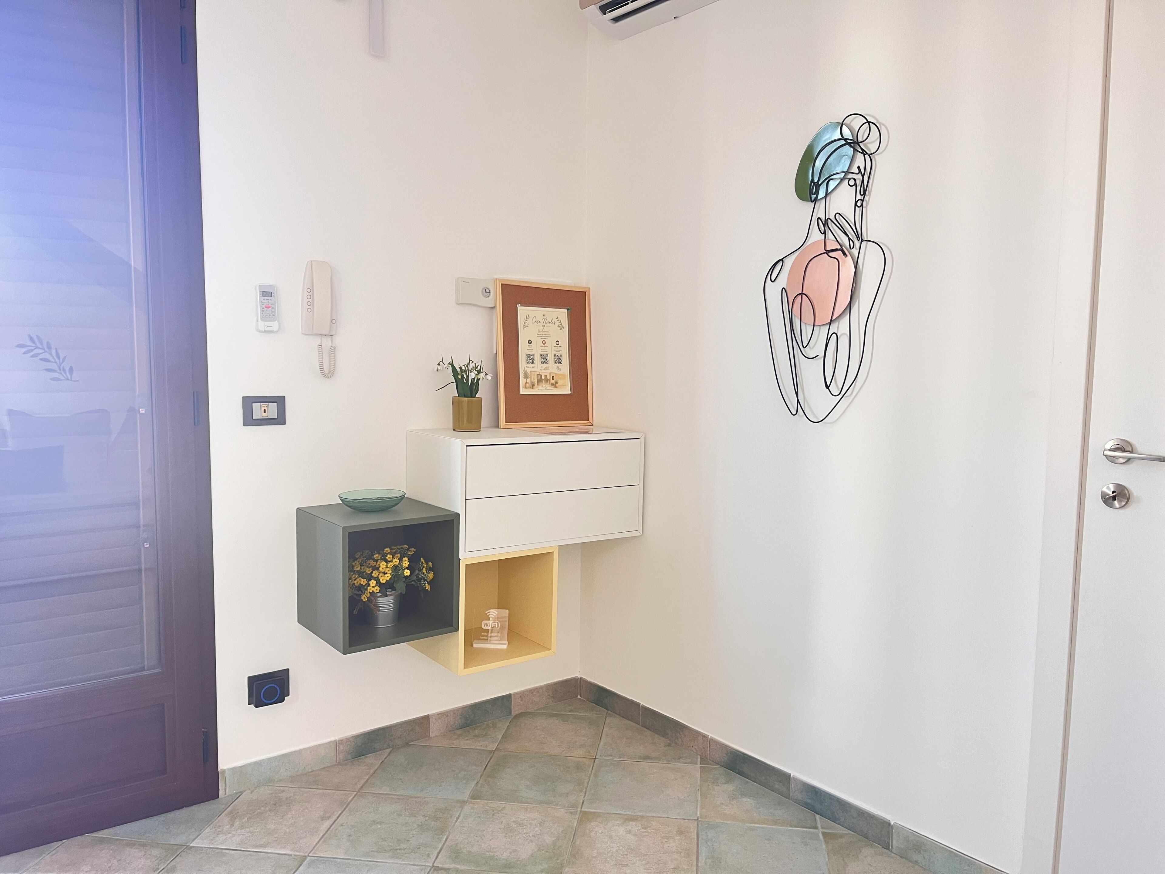 Un pasillo con una puerta, un teléfono sujeto a la pared, un certificado enmarcado, un pequeño estante con un cuenco y flores, y una obra de arte en la pared.